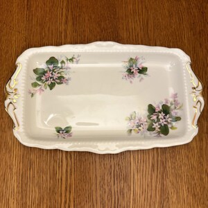 May include: A white rectangular serving tray with a gold trim and floral design. The tray has two handles on the sides.