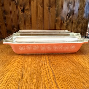 May include: A rectangular, coral-colored Pyrex baking dish with a clear glass lid. The dish features a white floral pattern along the side. The dish is sitting on a wooden surface.