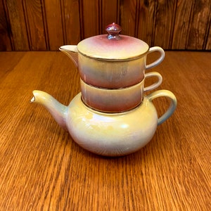 以下が含まれることがあります： 蓋と2つのカップが積み重ねられた、マルチカラーのセラミック製のティーポット。ティーポットは黄色、オレンジ、紫の釉薬がかかっています。カップは金色の縁取りが施された白です。