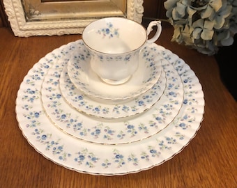 May include: A white porcelain tea set consisting of a teacup, saucer, and two plates, all featuring a delicate blue and purple floral pattern with scalloped edges and gold trim. The pieces are stacked neatly, showcasing the matching botanical design.