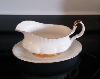 May include: White porcelain gravy boat with a gold rim and handle. The boat is on a matching saucer.