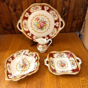 Może przedstawiać: Zestaw do herbaty z porcelany w stylu vintage, składający się z dużego półmiska, dzbanka do śmietanki i dwóch półmisków. Zestaw ma białe tło z kwiatowym wzorem i bordową obwódką ze złotymi wykończeniami.