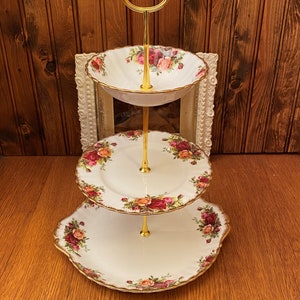 Puede incluir: Un soporte para tartas de tres niveles con un marco de metal dorado y platos de porcelana blanca decorados con un patrón floral. Los platos tienen un borde festoneado y presentan un borde dorado.