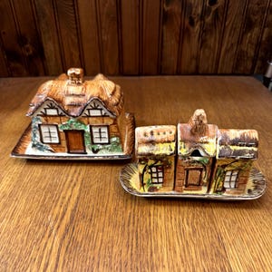 May include: A ceramic cottage-shaped butter dish with a thatched roof and a matching salt and pepper set. The butter dish and shakers are brown, white, and green, with a gold-coloured tray.