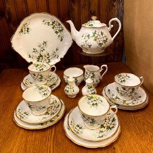 May include: A white tea set with a floral pattern of white flowers and green leaves. The set includes a teapot, creamer, sugar bowl, and four teacups and saucers.