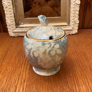 Peut inclure: Un pot à sucre en céramique blanche avec un motif floral bleu et blanc et une bordure dorée. Le couvercle est surmonté d'un petit bouton en forme de rose.