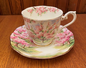 May include: A vintage teacup and saucer set with a floral pattern featuring pink and white blossoms on a cream background. The set has a gold trim around the edges.