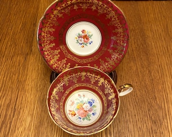 May include: A vintage teacup and saucer set with a red and gold floral pattern. The cup and saucer have a delicate, ornate design with a gold trim.