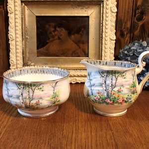 May include: A vintage porcelain sugar bowl and creamer set with a delicate floral and birch tree design. The set features a white background with a light blue sky and a lake scene. The trim is gold and gray.