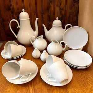 Peut inclure: Un ensemble de vaisselle en céramique blanche, comprenant des théières, des crémier, des tasses, des soucoupes et des assiettes. Les pièces ont une surface texturée et sont disposées sur une table en bois. L'ensemble comprend deux théières, deux crémiers, deux salières et poivrières et plusieurs tasses et assiettes.