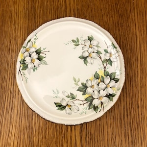 Puede incluir: Plato de cerámica blanco con borde dorado y un diseño floral con flores blancas con centros amarillos y hojas verdes.