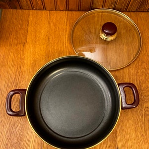 May include: A black pot with a gold rim and two red handles. The pot has a glass lid with a gold rim and a brown knob.