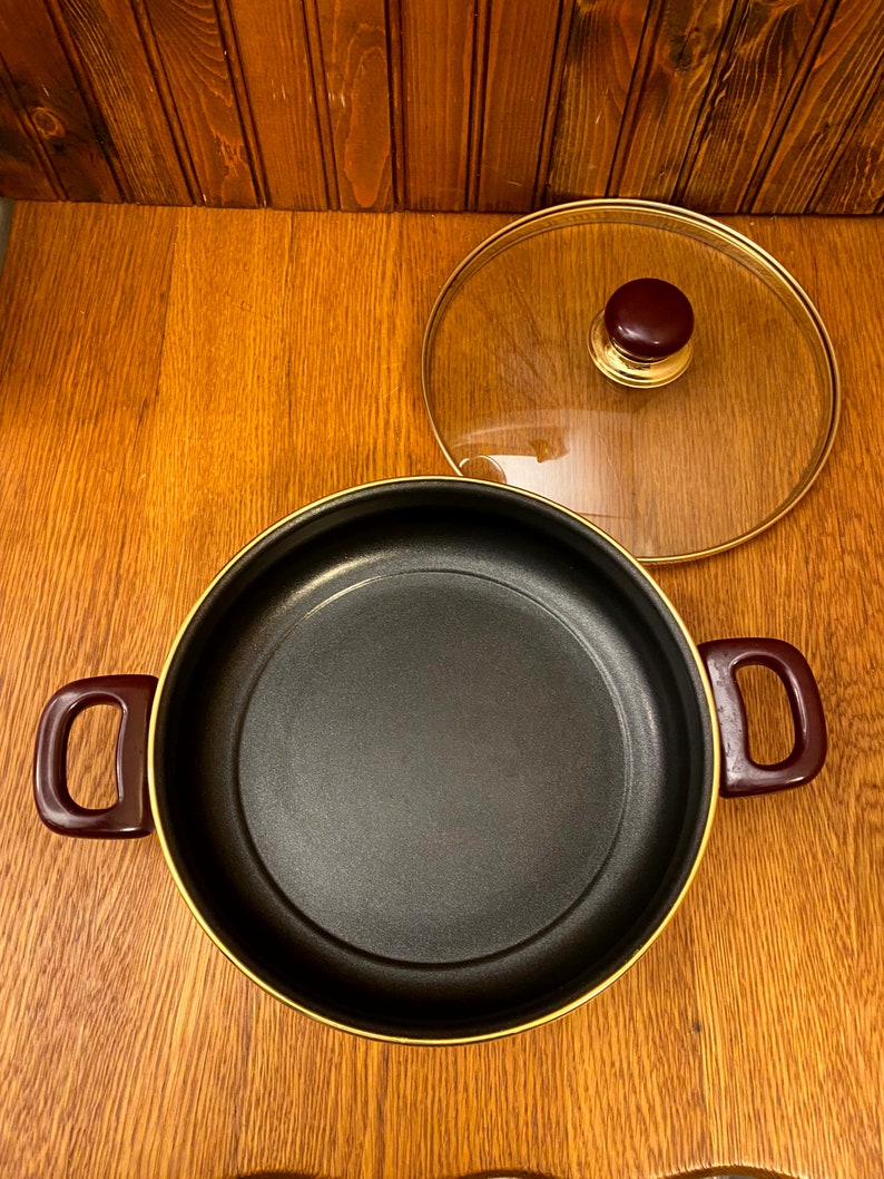 May include: A black pot with a gold rim and two red handles. The pot has a glass lid with a gold rim and a brown knob.