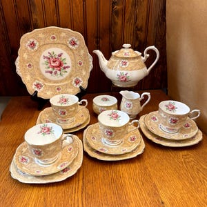 Może przedstawiać: Zestaw do herbaty w stylu vintage z kwiatowym wzorem. Zestaw zawiera czajnik, dzbanek do śmietanki, cukiernicę i cztery filiżanki do herbaty i spodeczki. Filiżanki do herbaty i spodeczki mają kwadratowy kształt.