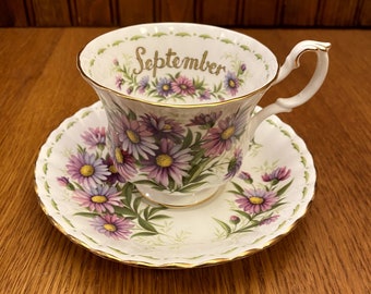 May include: A white teacup and saucer with a floral pattern and gold trim. The teacup has the word "September" written on it.