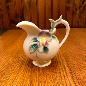 May include: A cream-coloured ceramic creamer with a floral design. The creamer has a round body, a curved handle, and a spout. The floral design includes purple, yellow, and pink flowers with green leaves. It sits on a small, round base.
