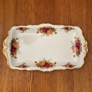 Puede incluir: Bandeja rectangular blanca con borde dorado y estampado floral de rosas. La bandeja presenta racimos de rosas rojas y amarillas con hojas verdes. Los bordes tienen un diseño festoneado.