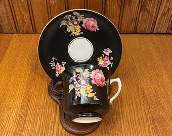 May include: A black teacup and saucer set with floral designs in pink, purple, and yellow. The cup and saucer are displayed on a dark wooden stand, set against a wooden background. The teacup has a white interior and handle.
