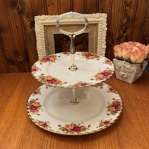 Puede incluir: Un soporte para tartas de porcelana blanca de dos niveles con un asa plateada. El soporte está decorado con un patrón floral de rosas rojas, rosas y amarillas. El soporte tiene un borde dorado.