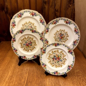Peut inclure: Ensemble de quatre assiettes en céramique blanche avec un motif floral. Les assiettes ont un bord doré et un motif floral doré au centre. Les assiettes sont disposées en pile, la plus grande assiette en bas et la plus petite assiette en haut.