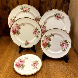 May include: A set of six white porcelain plates with a pink rose floral design and gold trim. The plates are displayed on black stands against a wooden background. The plates vary in size.