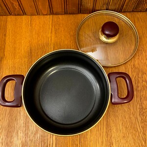 May include: A black pot with a gold rim and burgundy handles. The pot has a glass lid with a gold rim and a burgundy knob.