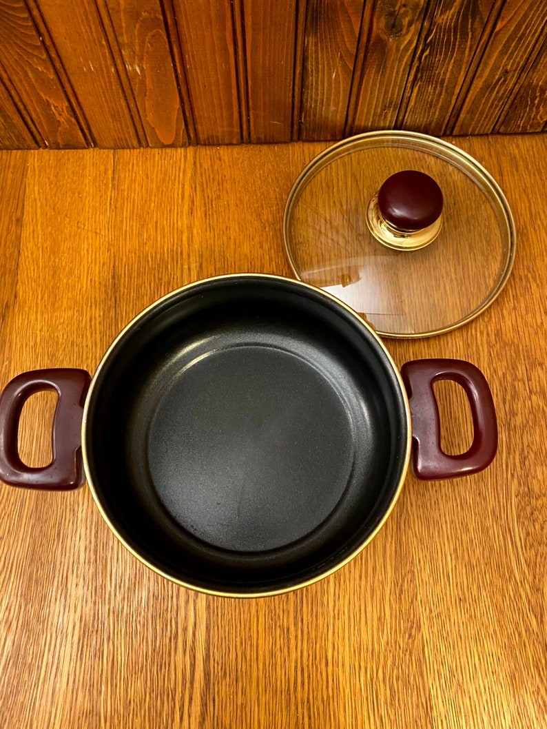 May include: A black pot with a gold rim and burgundy handles. The pot has a glass lid with a gold rim and a burgundy knob.