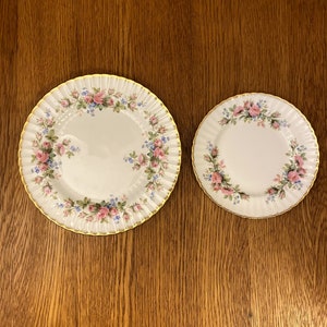 May include: Two white porcelain plates with a floral pattern of pink and blue flowers. The larger plate has a gold rim and the smaller plate has a gold rim.
