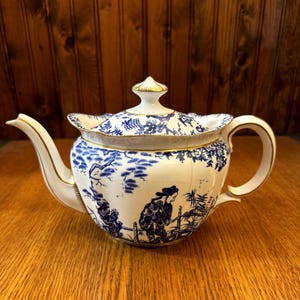 May include: A white porcelain teapot featuring a detailed blue transferware pattern of a figure in a landscape with trees and bamboo. The teapot has a scalloped lid with a gold-trimmed finial, a curved spout, and a gold-accented handle.