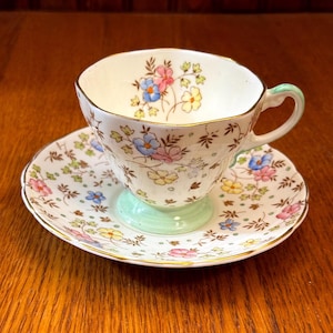 May include: A vintage teacup and saucer set. The white porcelain cup and saucer are decorated with colorful floral patterns. The cup has a light green handle and base, and the edges are trimmed with gold. The saucer also has a gold trim.