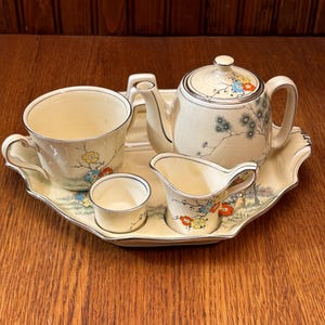 May include: A vintage tea set in cream, including a teapot, teacup, creamer, sugar bowl, and tray. The set is adorned with floral and tree branch designs in blue, yellow, and red. The tray is approximately 30 cm long.