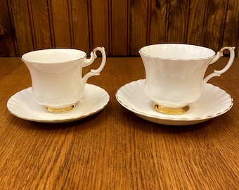 May include: Two white teacups and saucers with gold trim. The cups have a fluted design and the saucers have a scalloped edge.