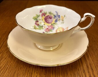 May include: A white teacup and saucer set with a floral design. The cup has a gold rim and the saucer has a pink rim.