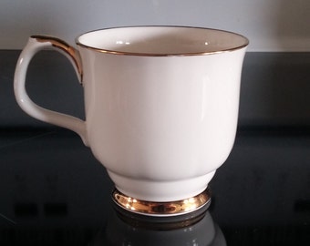 May include: A white ceramic mug with a gold rim and base. The mug has a rounded shape and a handle.