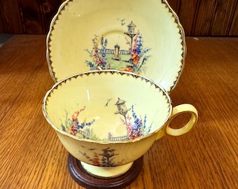May include: A vintage yellow teacup and saucer set. The cup and saucer feature a decorative floral and birdhouse design with a scalloped edge. The teacup sits on a small, dark wooden base. The set is likely made of porcelain.