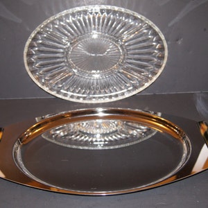 Puede incluir: Una bandeja de servir ovalada de plata con un asa de caparazón de tortuga. La bandeja tiene una superficie de espejo y está colocada encima de una fuente de servir de vidrio transparente con un patrón decorativo.