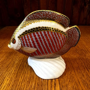 Puede incluir: Figura decorativa de pez de cerámica. El pez presenta un cuerpo blanco con detalles a rayas rojas y grises, acentuado con adornos dorados. Descansa sobre una base de concha blanca. El pez tiene un diseño detallado y es probablemente un artículo de colección.