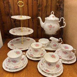 Może przedstawiać: Biały zestaw do herbaty z kwiatowym wzorem. Zestaw zawiera czajnik, dzbanek do śmietanki, cukiernicę i cztery filiżanki do herbaty z talerzykami. Filiżanki do herbaty i talerzyki mają delikatny kwiatowy wzór z różowymi różami i szarymi liśćmi. Zestaw jest prezentowany na drewnianym stole z trójstopniowym stojakiem na ciasto.