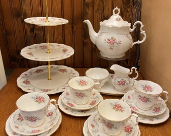 Op de afbeelding: Een wit theeservies met een bloemenpatroon. De set bevat een theepot, een roomkan, een suikerpot en vier theekopjes met schoteltjes. De theekopjes en schoteltjes hebben een delicaat bloemenpatroon met roze rozen en grijze bladeren. De set wordt gepresenteerd op een houten tafel met een drielaags taartstandaard.