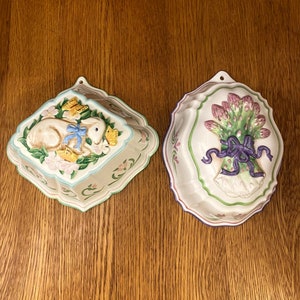 Peut inclure: Deux moules à gâteau en céramique avec des boucles de suspension. Un moule représente un lapin blanc avec un ruban bleu et des papillons jaunes. L'autre moule représente un bouquet de fleurs violettes avec une tige verte et un ruban violet.