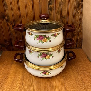 Könnte beinhalten: Ein Satz aus drei weißen Emaille-Töpfen mit goldenen Verzierungen und einem Blumenmuster. Die Töpfe haben rote Griffe und Deckel mit Glasoberflächen.