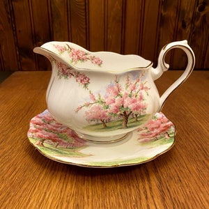 May include: A white porcelain gravy boat and saucer with a floral design featuring pink and white blossoms on a tree. The design is reminiscent of a spring garden.