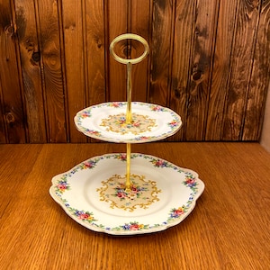 Peut inclure: Un support à gâteau à deux étages avec un support en métal doré et des assiettes en porcelaine blanche avec des motifs floraux. Les assiettes ont un bord festonné et une bordure dorée.