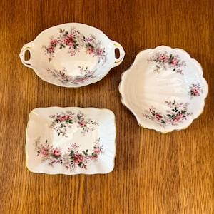 Peut inclure: Ensemble de trois pièces de vaisselle en porcelaine vintage. L'ensemble comprend un plat ovale avec des poignées, un plat à bord festonné et un plat rectangulaire. Chaque pièce est blanche avec un motif floral de roses roses et une bordure dorée.