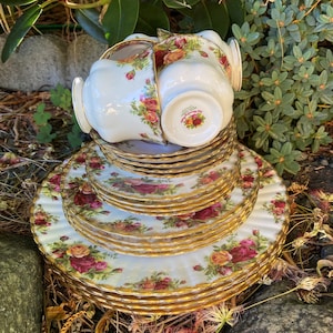 Puede incluir: Un juego vintage de tazas y platillos de té con un patrón floral y ribete dorado. Las tazas y los platillos están apilados uno encima del otro y están rodeados de follaje verde.
