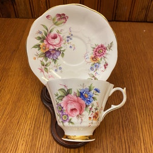 Puede incluir: Una taza de té y un platillo blancos con un patrón floral en tonos de rosa, morado y amarillo. La taza y el platillo tienen un borde dorado.