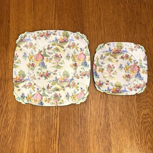 Può includere: Due piatti in ceramica vintage con motivi floreali. Il piatto più grande ha un bordo verde e quello più piccolo ha un bordo verde e bianco. Entrambi i piatti presentano un design cottagecore con fiori, case e altri dettagli.