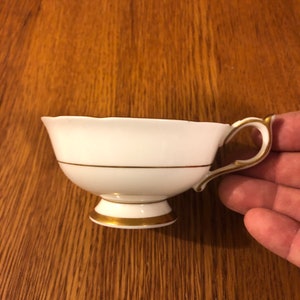 May include: A white teacup with a gold rim and a gold band around the middle. The teacup has a scalloped edge and a small handle.
