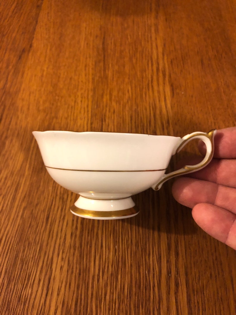 May include: A white teacup with a gold rim and a gold band around the middle. The teacup has a scalloped edge and a small handle.