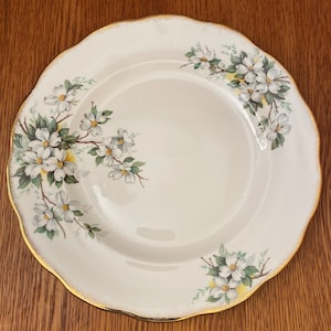 Puede incluir: Un plato de cena de cerámica blanca con un borde dorado y un diseño floral con flores blancas y hojas verdes.